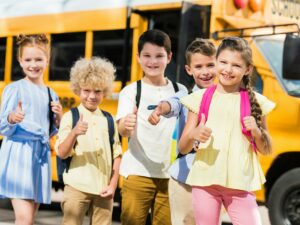 group of happy pupils looking at camera and showing thumbs up in front of school bus