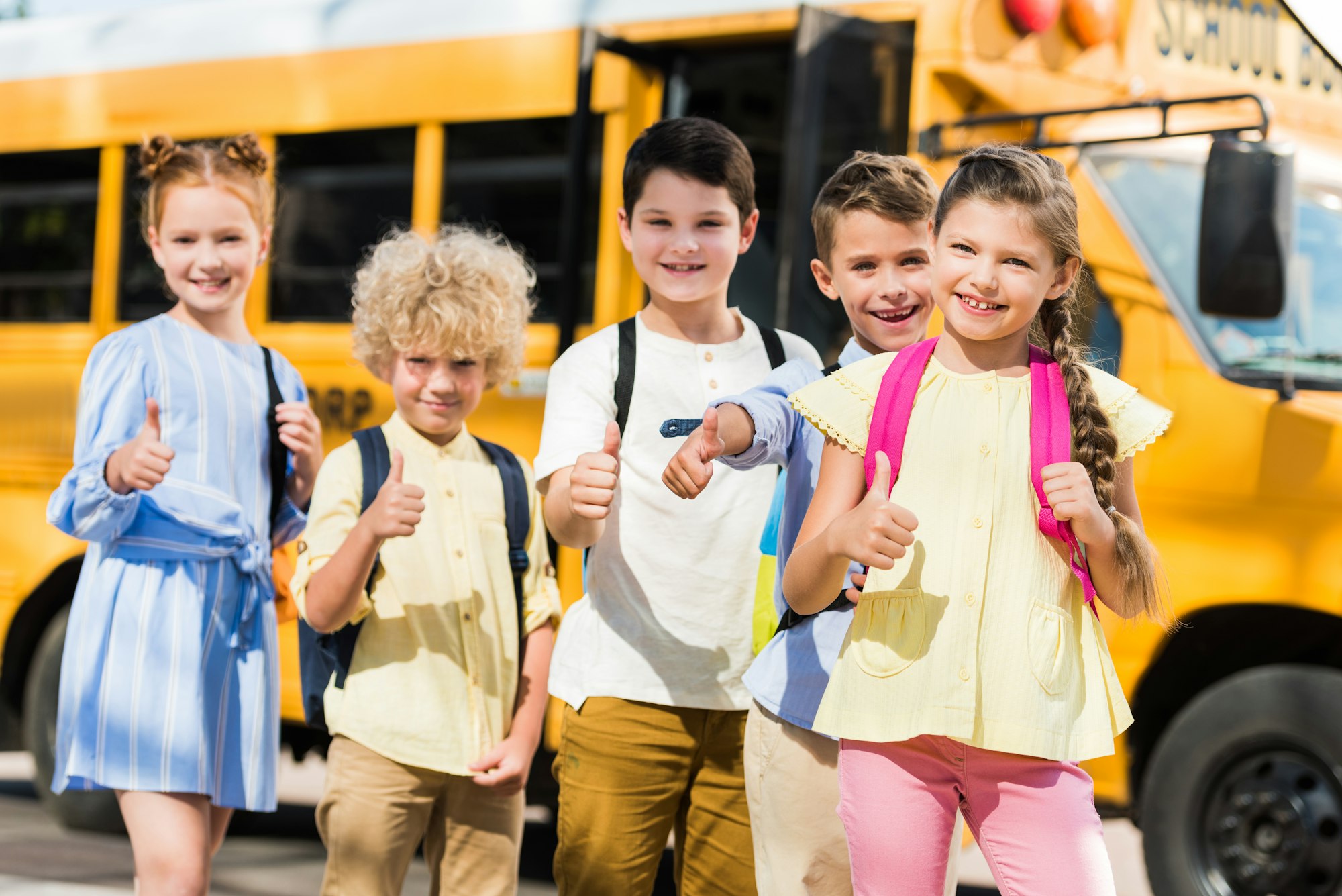 group of happy pupils looking at camera and showing thumbs up in front of school bus